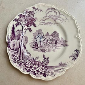 May include: A cream-coloured ceramic plate with a scalloped edge featuring a purple transferware print. The design depicts a landscape with a house, a body of water, trees, and floral motifs. The intricate scene covers the centre and rim.