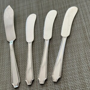 May include: Four silver butter spreaders with curved blades and decorative handles are arranged on a textured grey surface. The spreaders have a classic design, suitable for serving butter or other spreads at a dining table.