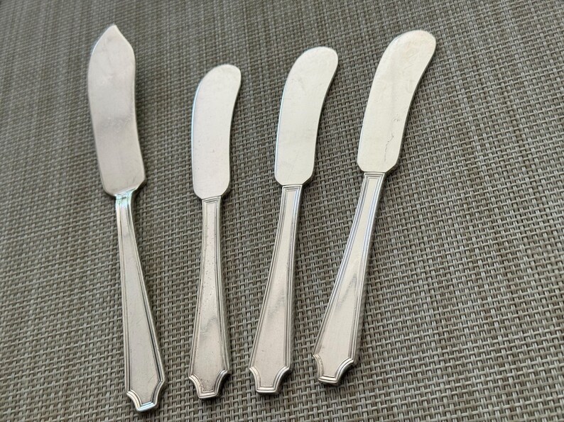 May include: Four silver butter spreaders with curved blades and decorative handles are arranged on a textured grey surface. The spreaders have a classic design, suitable for serving butter or other spreads at a dining table.