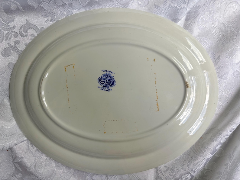 May include: An oval-shaped white ceramic serving platter with a smooth, glossy finish. The underside features a blue printed maker's mark that reads Willow Wood & Sons England. There are two faint, rectangular orange-toned marks on the surface.