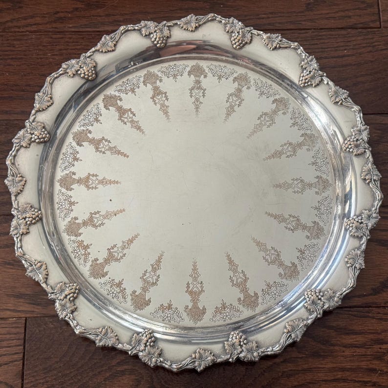 May include: A round silver-toned serving tray featuring an ornate, raised border decorated with embossed grape clusters and vine leaves. The center of the tray displays a radial pattern of etched, gold-toned floral and scrollwork motifs.