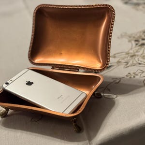 May include: A small, rectangular copper trinket box with a hinged lid, resting on ornate feet. Inside, a silver smartphone with a black Apple logo is visible. The box has a decorative, textured border and a polished finish.