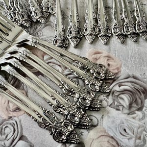 May include: A collection of ornate silver forks and serving utensils. The silverware features detailed floral and scrollwork designs. The pieces are arranged on a floral patterned fabric, creating a vintage aesthetic.