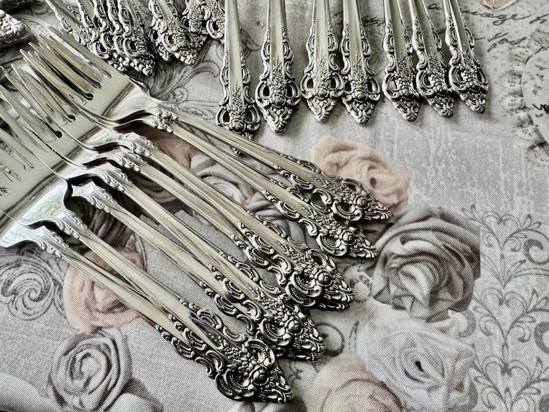 May include: A collection of ornate silver forks and serving utensils. The silverware features detailed floral and scrollwork designs. The pieces are arranged on a floral patterned fabric, creating a vintage aesthetic.