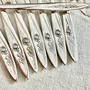 Op de afbeelding: Close-up van een set zilverkleurig bestek. Elk stuk heeft een rozenontwerp op het handvat. Het bestek is gerangschikt op een getextureerd, lichtgekleurd oppervlak, wat wijst op een vintage of antieke esthetiek.
