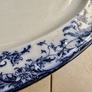 Puede incluir: Primer plano de un plato de cer&aacute;mica blanco con un dise&ntilde;o floral azul alrededor del borde. El patr&oacute;n presenta flores y follaje estilizados. El plato tiene una forma ligeramente irregular y un acabado brillante.