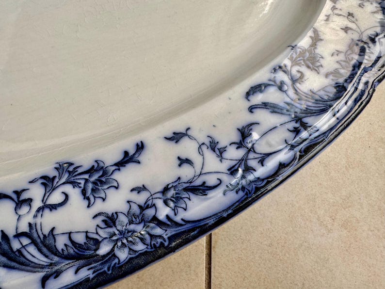 Puede incluir: Primer plano de un plato de cer&aacute;mica blanco con un dise&ntilde;o floral azul alrededor del borde. El patr&oacute;n presenta flores y follaje estilizados. El plato tiene una forma ligeramente irregular y un acabado brillante.