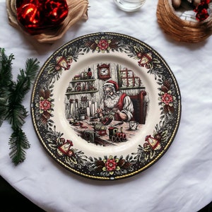 May include: A decorative plate featuring a detailed illustration of Santa Claus in his workshop. The plate has a cream-colored center with a black and gold border adorned with floral and bell motifs. Santa is depicted crafting toys.