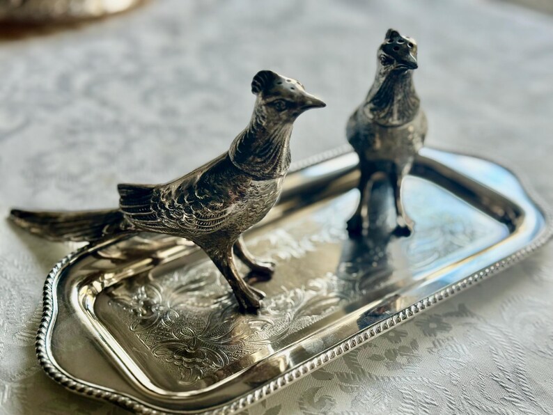 May include: A silver-toned metal serving tray with two bird-shaped salt and pepper shakers. The tray has a decorative floral pattern and a beaded edge. The birds are detailed with feathers and stand on the tray.