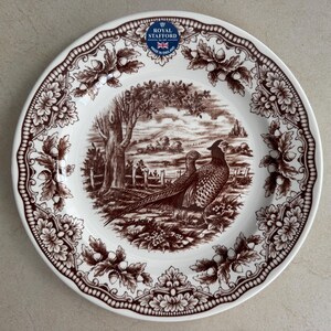 Op de afbeelding: Een wit keramieken bord met een bruin transferware patroon van twee fazanten in een landschap met bomen en een hek. De rand toont een ingewikkeld ontwerp van eikenbladeren en eikels. Het Royal Stafford Made in England logo is zichtbaar.