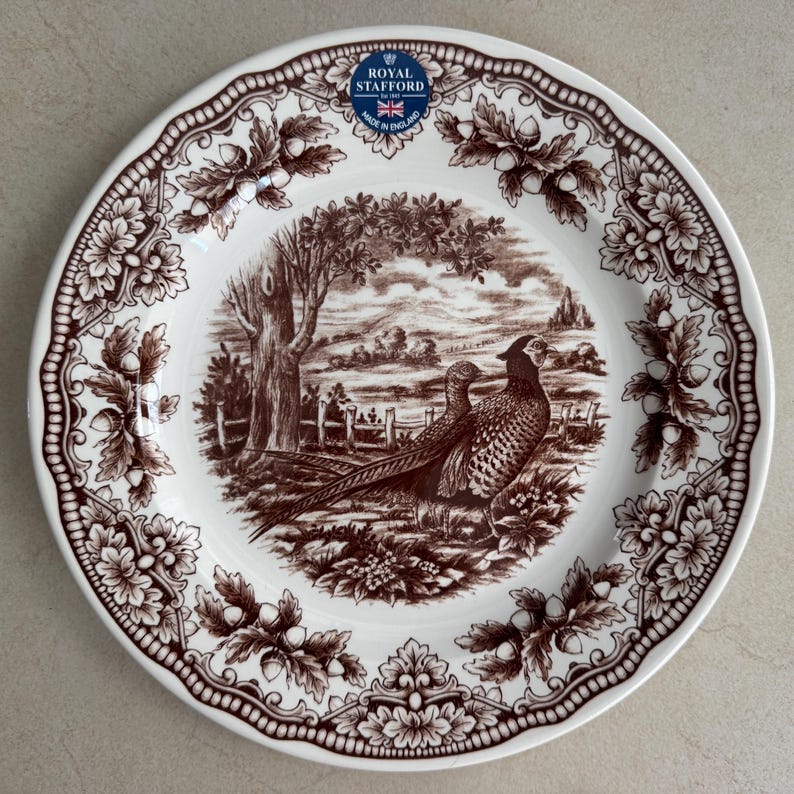 Op de afbeelding: Een wit keramieken bord met een bruin transferware patroon van twee fazanten in een landschap met bomen en een hek. De rand toont een ingewikkeld ontwerp van eikenbladeren en eikels. Het Royal Stafford Made in England logo is zichtbaar.