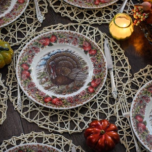 K&ouml;nnte beinhalten: Eine Thanksgiving-Tischdekoration mit wei&szlig;en Tellern, die ein Truthahn-Design zeigen, umgeben von geflochtenen Tischsets und dekorativen K&uuml;rbissen.