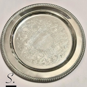 May include: A round silver-colored metal serving tray featuring a smooth, plain center surrounded by an intricate, embossed floral and scrollwork pattern. The outer edge of the tray is finished with a raised, twisted rope-style border design.