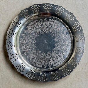 May include: A round silver-toned metal tray featuring an intricate embossed floral and scrollwork pattern in the center. The wide, scalloped rim has a decorative pierced cutout design that encircles the entire edge of the tray.