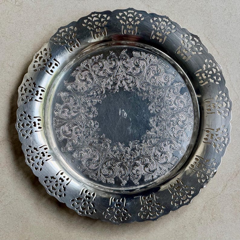 May include: A round silver-toned metal tray featuring an intricate embossed floral and scrollwork pattern in the center. The wide, scalloped rim has a decorative pierced cutout design that encircles the entire edge of the tray.