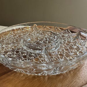 May include: Clear glass serving dish with a starburst pattern. The dish has a central dip bowl and is divided into sections. It sits on a wooden tray, perfect for serving appetizers or snacks.