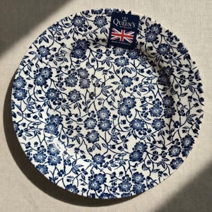 May include: A round ceramic plate featuring a dark blue floral pattern on a white background. A small rectangular tag is attached to the rim, displaying the text Queen's by Churchill, a Union Jack flag, and the words Made in England.