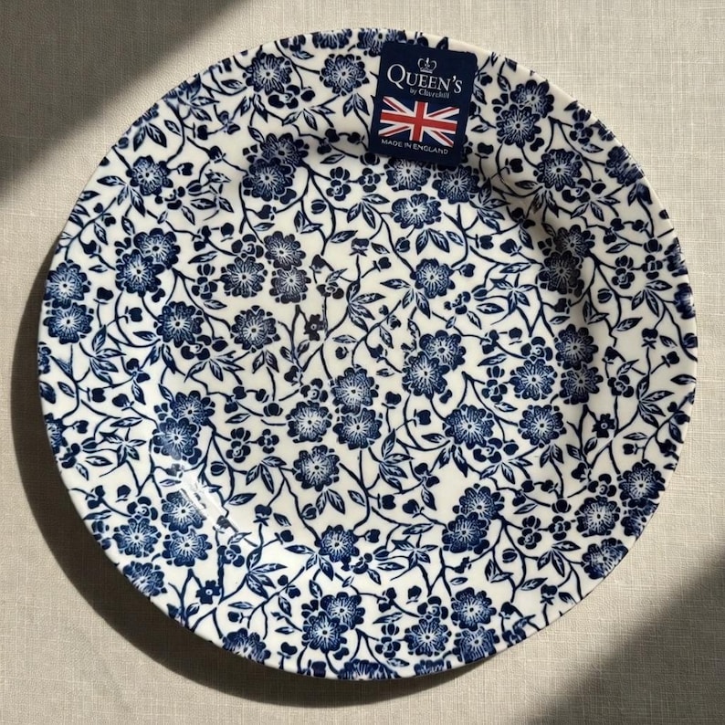 May include: A round ceramic plate featuring a dark blue floral pattern on a white background. A small rectangular tag is attached to the rim, displaying the text Queen's by Churchill, a Union Jack flag, and the words Made in England.