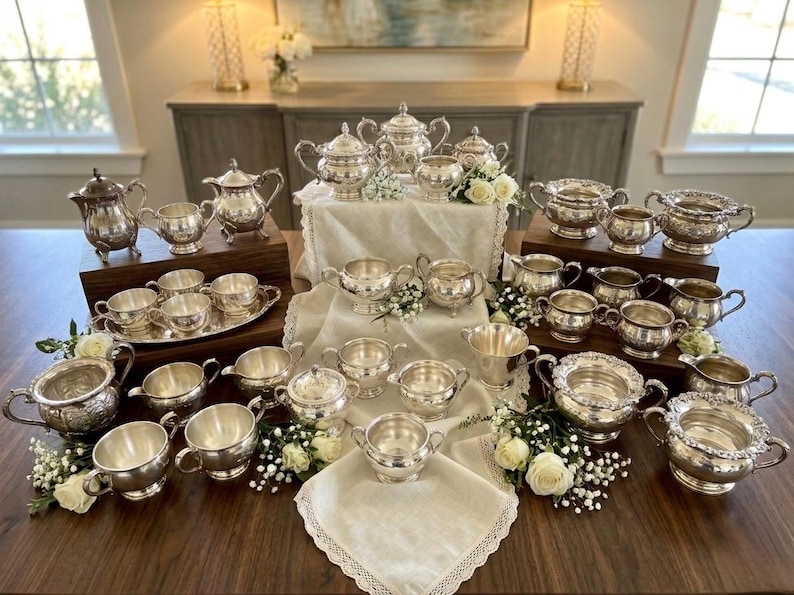 Puede incluir: Una gran colecci&oacute;n de piezas de servicio de t&eacute; plateadas, que incluye varias teteras, azucareros, cremeras y tazas. Los art&iacute;culos presentan detalles en relieve ornamentados, asas curvas y superficies met&aacute;licas pulidas, dispuestos sobre soportes de madera.