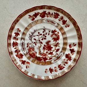 May include: A round ceramic saucer with a scalloped edge, featuring a white base decorated with deep red floral branches and a gold geometric border pattern. The center displays a detailed botanical motif with red blossoms and brown stems.