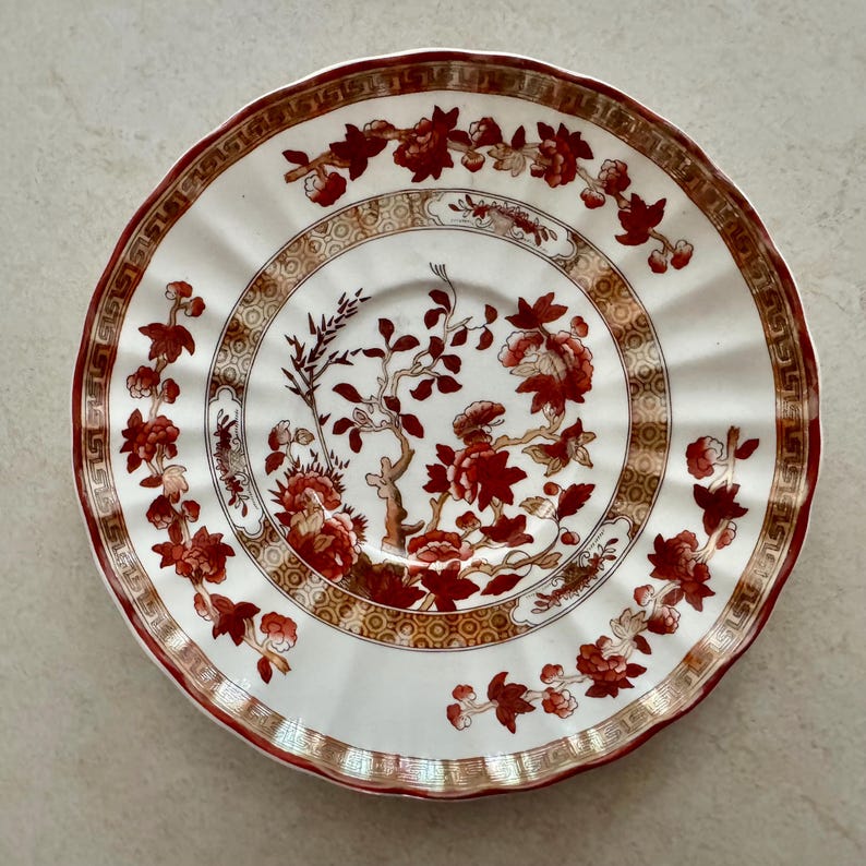 May include: A round ceramic saucer with a scalloped edge, featuring a white base decorated with deep red floral branches and a gold geometric border pattern. The center displays a detailed botanical motif with red blossoms and brown stems.
