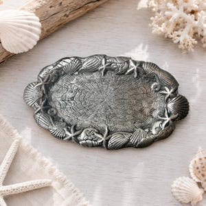 May include: An oval-shaped metal tray with a dark, antique silver finish. The center features a textured, radiating fan pattern, while the raised border is decorated with a relief design of various seashells and starfish arranged around the edge.
