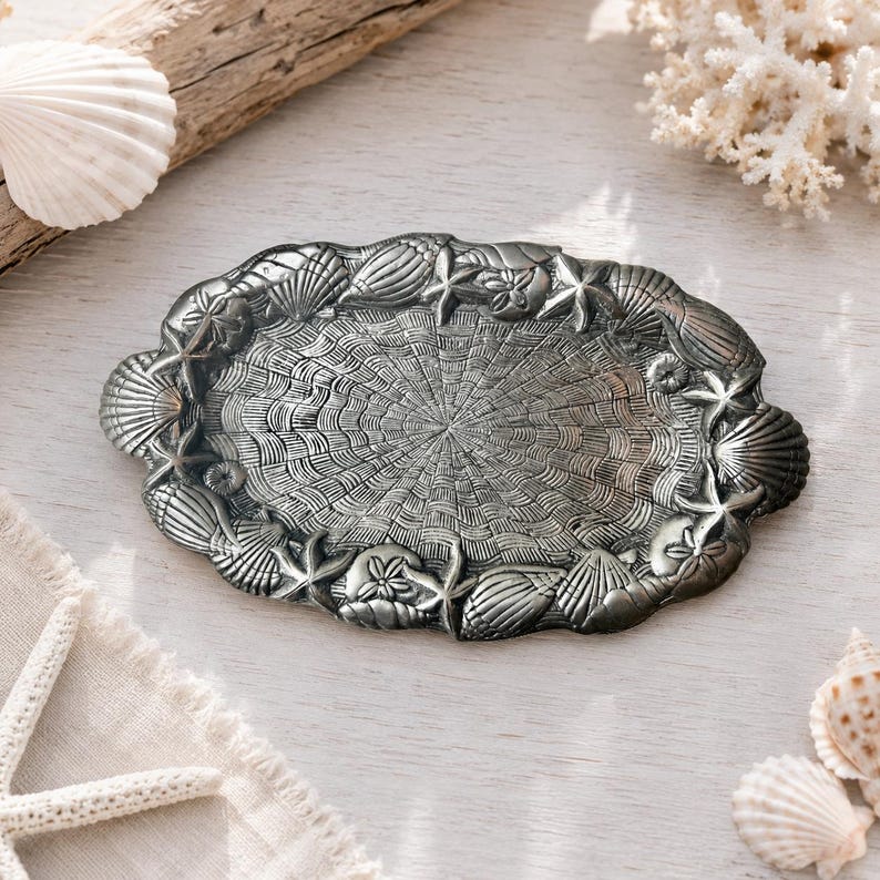 May include: An oval-shaped metal tray with a dark, antique silver finish. The center features a textured, radiating fan pattern, while the raised border is decorated with a relief design of various seashells and starfish arranged around the edge.