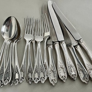 Puede incluir: Un conjunto de cubiertos plateados, que incluye cucharas, tenedores y cuchillos. Los mangos presentan un dise&ntilde;o floral ornamentado. Las cucharas est&aacute;n apiladas y los tenedores y cuchillos est&aacute;n dispuestos en fila, mostrando un estilo cl&aacute;sico y elegante.