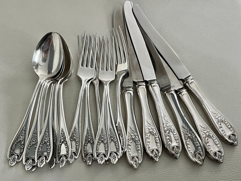 Puede incluir: Un conjunto de cubiertos plateados, que incluye cucharas, tenedores y cuchillos. Los mangos presentan un dise&ntilde;o floral ornamentado. Las cucharas est&aacute;n apiladas y los tenedores y cuchillos est&aacute;n dispuestos en fila, mostrando un estilo cl&aacute;sico y elegante.