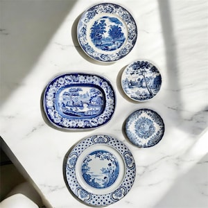 May include: An assortment of blue and white porcelain plates and a serving platter. The plates feature scenic landscape designs and floral patterns. The arrangement includes round and oval shapes, showcasing a classic aesthetic.