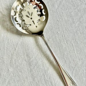 May include: A silver-colored slotted spoon with a round head featuring decorative cut-out patterns. The long handle has a rounded end with a small, engraved design. The spoon is designed for straining liquids and is likely used in cooking.