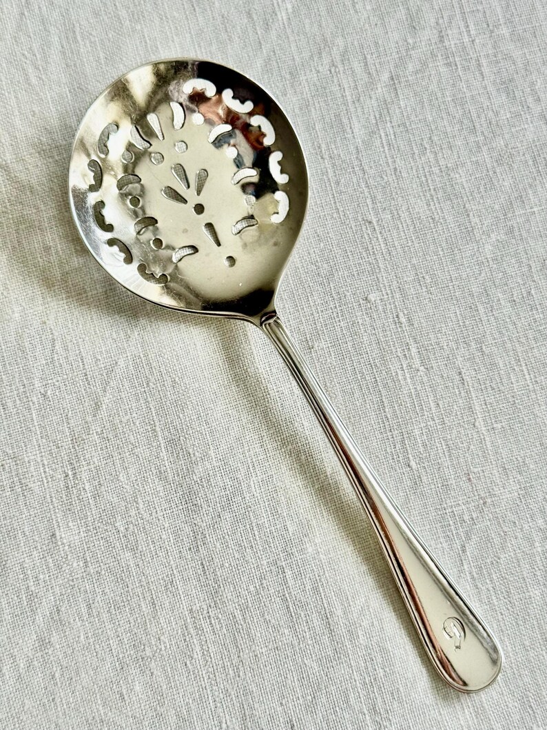 May include: A silver-colored slotted spoon with a round head featuring decorative cut-out patterns. The long handle has a rounded end with a small, engraved design. The spoon is designed for straining liquids and is likely used in cooking.