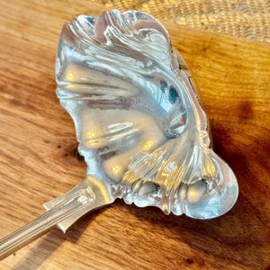 May include: A silver serving ladle with an ornate, decorative, and highly detailed design. The ladle has a long, slender handle and a wide, shallow bowl.