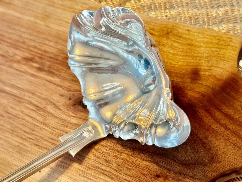 May include: A silver serving ladle with an ornate, decorative, and highly detailed design. The ladle has a long, slender handle and a wide, shallow bowl.