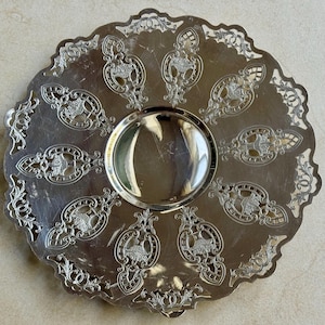 May include: A round silver-toned metal serving tray featuring a smooth, reflective center basin surrounded by a wide, ornate border. The border displays intricate cut-out patterns and embossed basket motifs arranged in a radial design.