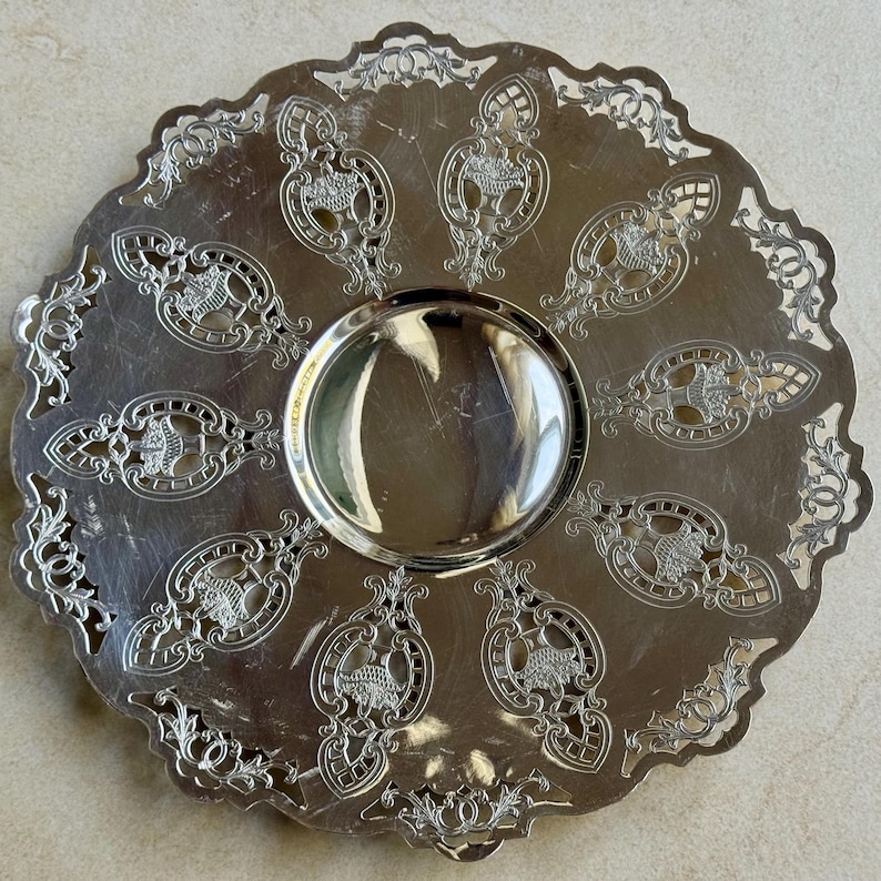May include: A round silver-toned metal serving tray featuring a smooth, reflective center basin surrounded by a wide, ornate border. The border displays intricate cut-out patterns and embossed basket motifs arranged in a radial design.