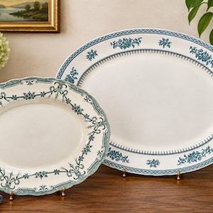 May include: Two oval ceramic serving platters in cream with teal blue floral and ribbon border patterns. The smaller platter features a scalloped edge, while the larger platter has a smooth rim with a geometric and floral design.
