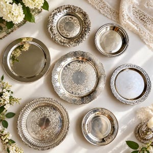 May include: A collection of six silver-toned metal plates and trays of varying sizes. The set includes three pieces with intricate embossed floral and scrollwork borders, two plain polished circular plates, and one smaller smooth plate.
