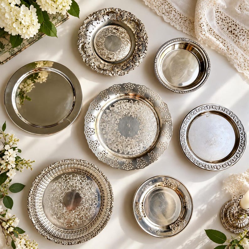 May include: A collection of six silver-toned metal plates and trays of varying sizes. The set includes three pieces with intricate embossed floral and scrollwork borders, two plain polished circular plates, and one smaller smooth plate.