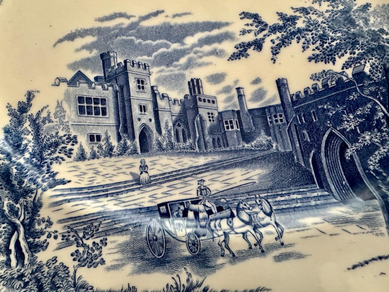 Op de afbeelding: Een cr&egrave;mekleurig keramieken bord met een blauwe transferprint van een groot stenen kasteel met torens en boogramen. Op de voorgrond rijdt een door paarden getrokken koets met een koetsier over een geplaveide binnenplaats.