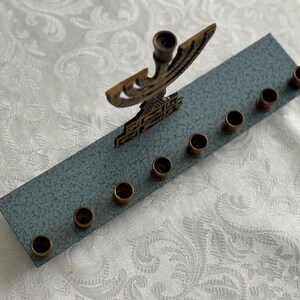 May include: A brass menorah with a decorative design, featuring a central holder and eight candle holders. The menorah is mounted on a rectangular, textured, blue-grey base. The background is a white, patterned fabric.