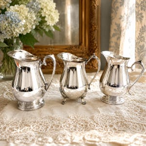 May include: Three polished silver pitchers with curved handles and pouring spouts. The left pitcher has a circular base, the center pitcher features three decorative feet, and the right pitcher stands on a circular pedestal base with a flared rim.