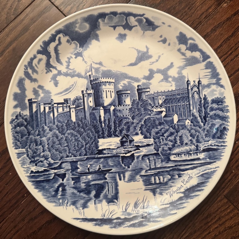 May include: A round ceramic plate featuring a blue and white transferware print of Windsor Castle. The scene depicts the castle architecture, surrounding trees, and a small boat on the water. The text Windsor Castle is printed on the lower right.