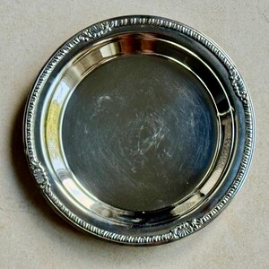 May include: A round silver-toned metal plate featuring a smooth, reflective center surface and a decorative border with a repeating ribbed pattern and small shell-like motifs at regular intervals along the outer rim.
