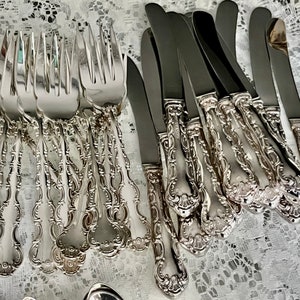May include: An assortment of ornate silver-colored flatware, including forks and knives, arranged on a white lace surface. The silverware features detailed, decorative handles with a floral design, suggesting a formal dining setting.