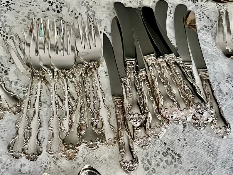 May include: An assortment of ornate silver-colored flatware, including forks and knives, arranged on a white lace surface. The silverware features detailed, decorative handles with a floral design, suggesting a formal dining setting.