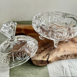 May include: A clear glass candy dish with a matching lid, featuring a faceted geometric pattern and starburst motifs. The bowl is supported by three small feet, and the lid includes a decorative handle with a scalloped top edge.