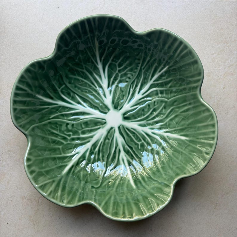 May include: A ceramic bowl shaped like a cabbage leaf, featuring a glossy finish in shades of green. The design includes a central point with white, raised veins radiating outward toward the scalloped, wavy edges of the bowl.
