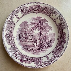 May include: A round ceramic plate featuring a purple transferware pattern on a cream background. The centre depicts a rural landscape with a cottage, a person on horseback, and dogs. The rim is decorated with a detailed floral and agricultural motif.