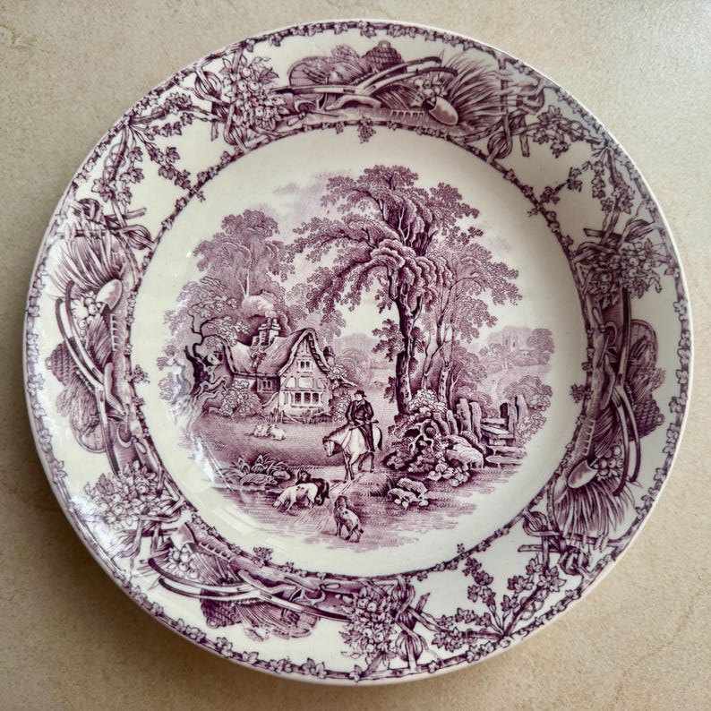 May include: A round ceramic plate featuring a purple transferware pattern on a cream background. The centre depicts a rural landscape with a cottage, a person on horseback, and dogs. The rim is decorated with a detailed floral and agricultural motif.